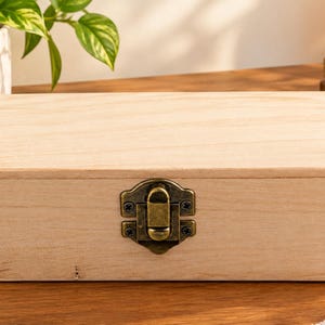 May include: A rectangular light-colored wooden box with a smooth, unfinished surface. The box features a hinged lid and a central antique brass-toned metal latch mechanism secured with small screws on the front panel for closure.