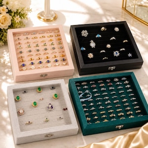 May include: Four square jewelry display cases with glass lids, featuring velvet-lined interiors in light pink, black, light gray, and dark teal. Each case contains multiple rows of slots filled with various rings, including gold, silver, and gemstone designs.