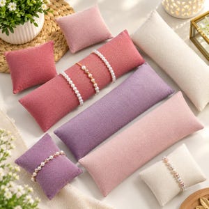 May include: A collection of rectangular and square fabric display pillows in shades of pink, purple, and cream. Several pillows feature beaded bracelets draped across their surfaces. The pillows have a textured linen-like finish and are arranged in a group.