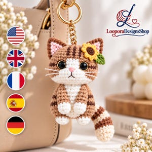 Può includere: Un portachiavi a forma di gatto lavorato all'uncinetto nei colori marrone e bianco, con un accessorio a girasole sulla testa, grandi occhi neri, naso rosa e un anello in metallo dorato. Il design presenta strisce marrone scuro su testa e coda.