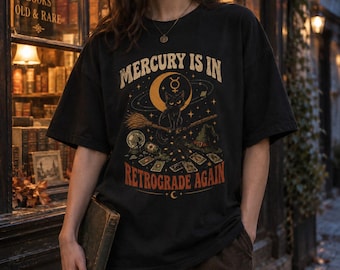 May include: A black short-sleeve t-shirt featuring a graphic print of a cat sitting on a broomstick under a crescent moon and the text Mercury is in Retrograde Again. The design includes tarot cards, a crystal ball, and a witch hat in orange.