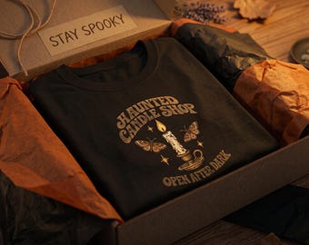 May include: A black t-shirt inside a cardboard box with the text STAY SPOOKY on the inner lid. The shirt features a gold graphic of a lit candle in a holder, two moths, and the text HAUNTED CANDLE SHOP and OPEN AFTER DARK printed in gold.