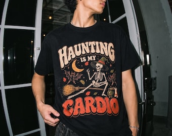 May include: A black short-sleeve t-shirt featuring a graphic print of a skeleton wearing a witch hat and riding a broomstick. The text reads "HAUNTING IS MY CARDIO" in bold, stylized lettering, accompanied by illustrations of bats, a crescent moon, and a cauldron.