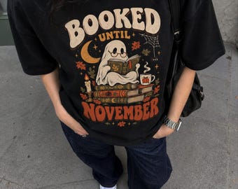May include: A black t-shirt featuring a graphic print of a ghost wearing glasses and reading a book while sitting on a stack of books. The text reads BOOKED UNTIL NOVEMBER. The design includes a crescent moon, a candle, a mug, and autumn leaves.