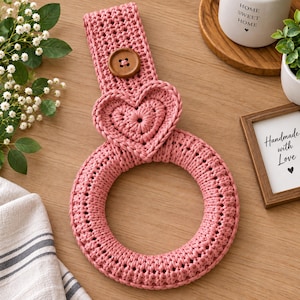 May include: A dusty rose crochet towel holder featuring a circular ring base, a heart-shaped accent, and a vertical strap secured with a single brown wooden button. The textile construction displays a consistent knit pattern throughout the design.