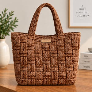 May include: A brown tote bag featuring a woven basket-weave pattern made from thick cord. The bag has two sturdy top handles and a small rectangular leather-like tag centered on the front with the word HANDMADE embossed in black capital letters.