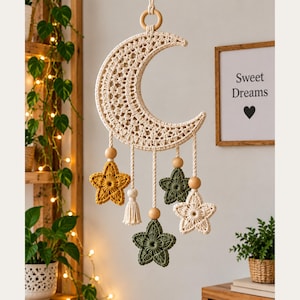 Puede incluir: Un tapiz de macramé color crema con forma de media luna, que cuenta con un anillo de madera central para colgarlo. De la luna cuelgan cuatro estrellas de ganchillo en amarillo mostaza, verde oliva y crema, decoradas con cuentas de madera.