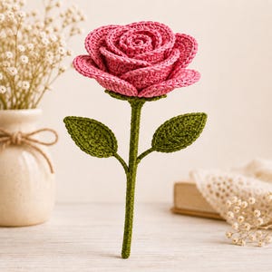 May include: A handcrafted crochet rose featuring a vibrant pink flower head with layered, textured petals. The stem is a solid dark green, complemented by two detailed, pointed green leaves extending from the sides of the central stalk.
