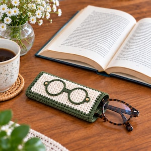 Peut inclure: Un étui à lunettes rectangulaire en crochet avec une bordure vert foncé et un panneau central crème. L'étui présente un contour brodé vert foncé représentant des lunettes rondes. Une paire de lunettes à motif écaille est rangée à l'intérieur.