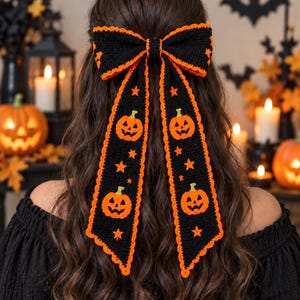 Puede incluir: Un gran lazo para el pelo de ganchillo negro con largas cintas colgantes. El lazo está ribeteado con hilo naranja brillante y decorado con pequeñas estrellas naranjas y motivos de calabazas de Halloween con tallos verdes.