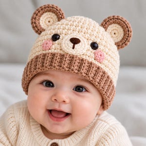 May include: A cream-colored crochet beanie featuring a brown ribbed brim and two round bear ears on top. The hat displays a bear face with a cream muzzle, a dark brown nose, two black button eyes, and two small pink circular cheeks on the sides.