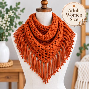 May include: A burnt orange crochet triangular scarf featuring an openwork diamond pattern and long fringe along the bottom edges. The scarf is displayed on a mannequin and includes a circular graphic with the text Adult Women Size.