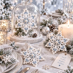 Peut inclure: Trois flocons de neige en crochet blanc et bleu clair avec des perles centrales. Un flocon est sous une cloche en verre, tandis que deux autres reposent sur une table à côté d'une étiquette portant l'inscription Small Stitches Brighter Holidays.