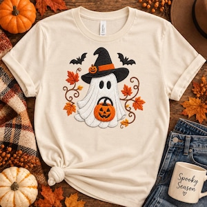 May include: A cream-colored short-sleeve t-shirt featuring an embroidered design of a white ghost wearing a black witch hat with an orange band and a small pumpkin. The ghost holds an orange jack-o'-lantern, surrounded by orange autumn leaves.