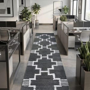 Puede incluir: Una alfombra de pasillo larga y rectangular con una base de color gris carbón oscuro y un patrón geométrico blanco repetitivo de formas escalonadas en forma de cruz. La alfombra está colocada en el centro sobre un suelo de hormigón.