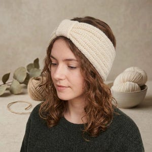 May include: A cream-colored knit headband featuring a ribbed texture and a central knotted detail. The headband is crafted from soft yarn and sits across the forehead, showcasing a consistent horizontal stitch pattern throughout the design.