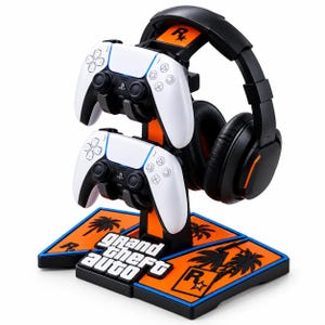 Puede incluir: Un soporte para accesorios de juego negro y naranja con el logotipo de Grand Theft Auto, gráficos de palmeras y la marca Rockstar Games. El soporte sostiene dos mandos de juego inalámbricos blancos y auriculares negros.
