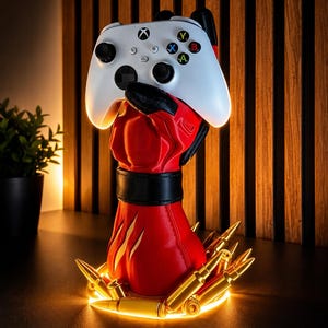May include: A white Xbox wireless controller held by a red, gloved hand sculpture. The base of the red hand is encircled by several gold-colored bullet casings, with a black band around the wrist. The entire display is illuminated from below.