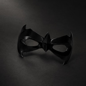 Könnte beinhalten: Eine einfarbig schwarze Augenmaske mit spitzem, kantigem Design und zwei ovalen Augenöffnungen. Die Maske hat eine glatte, glänzende Oberfläche und an den Seiten befinden sich scharfe, flügelartige Kanten, die eine symmetrische Form bilden.