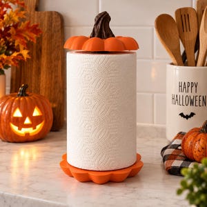 Puede incluir: Un soporte para papel de cocina con una base de color naranja brillante en forma de calabaza y una tapa superior a juego con forma de calabaza y tallo marrón oscuro. Está diseñado para sostener un rollo de papel blanco con textura.