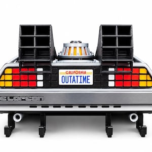Puede incluir: Un modelo de la parte trasera de un coche DeLorean plateado con rejillas negras, un componente de motor circular central y luces traseras rojas, amarillas y blancas. Una matrícula blanca de California con texto en rojo y azul dice OUTATIME.
