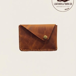 May include: A rectangular brown leather card holder with a triangular flap secured by a single brass-coloured metal snap button. The edges are stitched with thick thread. A circular logo in the corner reads LEATHER & YARN CO HANDMADE WITH LOVE.