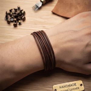 May include: A multi-strand dark brown leather bracelet worn on the wrist. Nearby are metal rivets, a leather crafting tool, a piece of leather, and a rectangular brass tag with the text HANDMADE GENUINE LEATHER engraved on the surface.
