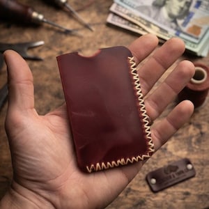 May include: A rectangular dark burgundy leather card holder featuring a smooth matte finish and a semi-circular cutout at the top edge. The side and bottom edges are joined with thick, light-coloured stitching in a visible cross-stitch pattern.