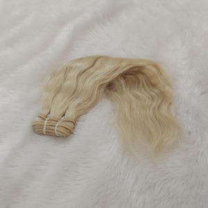 Puede incluir: Un paquete de extensiones de cabello rubio claro con textura ondulada. El cabello está sujeto en la parte superior con una banda de tejido horizontal, presentando un color pálido y platinado uniforme en toda la longitud de las hebras.