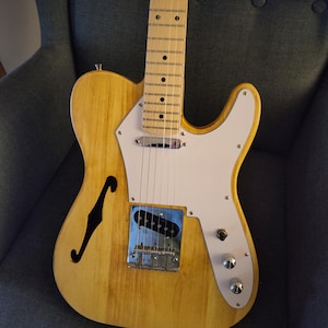Könnte beinhalten: Eine E-Gitarre mit hohlem Korpus in heller Naturholzoptik, weißem Schlagbrett und Ahorngriffbrett. Das Instrument verfügt über ein einzelnes F-Loch, verchromte Hardware, einen Steg-Tonabnehmer und zwei Regler im unteren Bereich des Korpus.