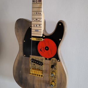 May include: An electric guitar with a weathered grey wooden body, a maple fretboard with decorative inlays, and a black pickguard featuring a circular red Polydor Visage vinyl record label. It includes gold hardware, an ornate bridge, and knobs.