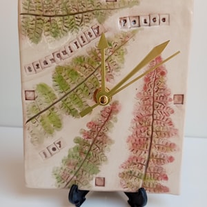 May include: A rectangular ceramic clock with a cream-coloured surface featuring embossed green and reddish-brown fern leaf patterns. The clock face includes the words tranquility, peace, and joy stamped into the clay, with gold-coloured hands.