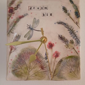 May include: A square ceramic wall clock with a cream-coloured surface featuring botanical illustrations of leaves, flowers, and a dragonfly. The text "dream big" is printed in black letters. The clock has three gold-coloured metal hands at the centre.