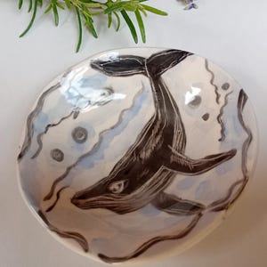 May include: A round ceramic dish featuring a hand-painted black whale illustration centred on a white background with light blue and grey abstract wavy lines and circular accents surrounding the whale, creating a detailed aquatic scene.