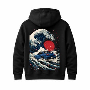 Könnte beinhalten: Ein schwarzer Kapuzenpullover mit einem Grafikdruck auf der Rückseite. Das Motiv zeigt einen blauen Sportwagen, der durch eine große, stilisierte Meereswelle mit weißem Schaum fährt, vor einem roten Sonnenkreis und rosa Kirschblüten.