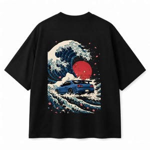 Op de afbeelding: Een zwart t-shirt met korte mouwen en een grafische print op de achterkant. Het ontwerp toont een blauwe sportwagen die door een grote gestileerde oceaangolf rijdt, met een opvallende rode zon op de achtergrond en verspreide roze bloesemblaadjes.
