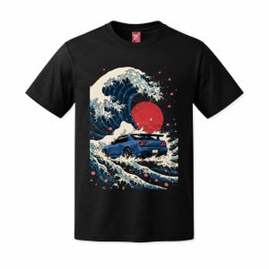 Może przedstawiać: Czarny t-shirt z krótkim rękawem z nadrukiem graficznym przedstawiającym niebieski samochód sportowy jadący przez dużą, stylizowaną falę oceaniczną. Wzór zawiera duże czerwone słońce w tle oraz rozrzucone różowe płatki kwiatów wiśni.