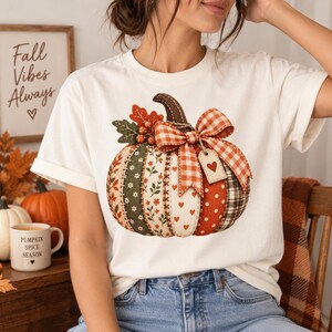May include: A cream-coloured t-shirt featuring a large graphic of a patchwork pumpkin. The pumpkin is decorated with various patterns including floral, polka dot, and plaid, topped with a large orange and white gingham bow and a small heart tag.
