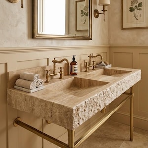 May include: A double vanity sink crafted from beige travertine stone with a rough, chiseled front edge. It features two brass faucets, a central soap dispenser, and a metal frame base. Three rolled white towels sit on a small tray on the left side.