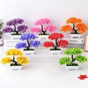 May include: A collection of seven artificial bonsai trees in white rectangular pots, featuring dark brown trunks and vibrant plastic foliage in purple, pink, orange, blue, red, yellow, and green colors, arranged on white surfaces.