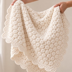 May include: A cream-coloured crochet blanket featuring a repeating pattern of small, raised floral motifs across the surface. The edges of the blanket are finished with a decorative scalloped border, creating a soft, textured appearance throughout.