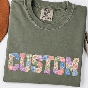 Może przedstawiać: T-shirt z okrągłym dekoltem w kolorze mchu z napisem CUSTOM na piersi w stylu patchworkowym. Litery mają różne pastelowe wzory, w tym kwiaty, kratkę vichy i kropki. Koszulka posiada metkę marki Comfort Colors.