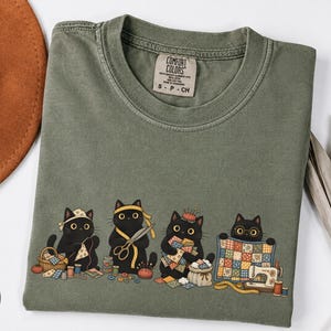 May include: A moss green t-shirt featuring a graphic of four black cats engaged in sewing and quilting activities. The cats are shown with sewing supplies including scissors, thread, a pincushion, and a sewing machine. The shirt has a Comfort Colors label.
