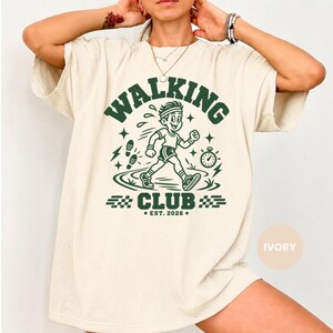 May include: An ivory short-sleeve t-shirt featuring a dark green graphic print. The design displays a person walking with the text WALKING CLUB and EST. 2026. The graphic includes a stopwatch, footprints, and checkered patterns.
