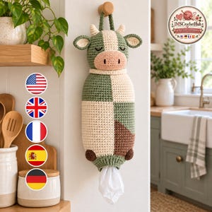 May include: A crochet cow-shaped plastic bag dispenser hanging on a wall hook. The dispenser features a cream, sage green, and brown color-blocked body with a pink snout and closed eyes. A white plastic bag protrudes from the bottom opening.