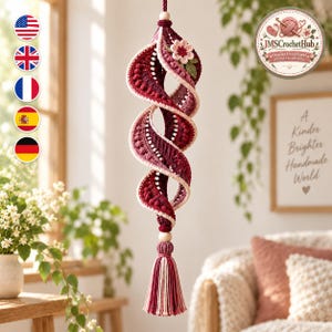 May include: A hanging spiral ornament crafted from crochet yarn in shades of burgundy, maroon, and cream. The structure features a textured, twisted design adorned with a small crocheted flower and green leaves at the top, finished with a tassel at the bottom.