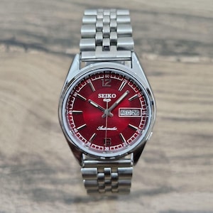 Op de afbeelding: Een automatisch Seiko 5 polshorloge met een levendige rode sunray-wijzerplaat met zilverkleurige wijzers en uurmarkeringen. Het horloge heeft een dag-datumvenster op de drie-uurpositie, Arabische cijfers op twaalf en zes, en een roestvrijstalen schakelarmband.