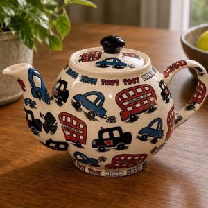 May include: A cream-colored ceramic teapot featuring a colorful pattern of red double-decker buses and blue cars. The design includes the text "TOOT TOOT" and "HONK HONK" printed in black and red. It has a black lid knob and a curved handle.