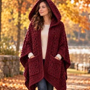 May include: A deep burgundy hooded crochet poncho featuring a chunky knit texture, an open front design, and two large square patch pockets on the front. The garment is worn over a plain white long-sleeved top, dark blue denim jeans, and brown boots.
