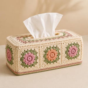May include: A rectangular tissue box cover crafted from cream-colored yarn with a granny square pattern. Each square features a central flower motif in shades of dusty pink or muted orange, surrounded by sage green petals and a cream border.